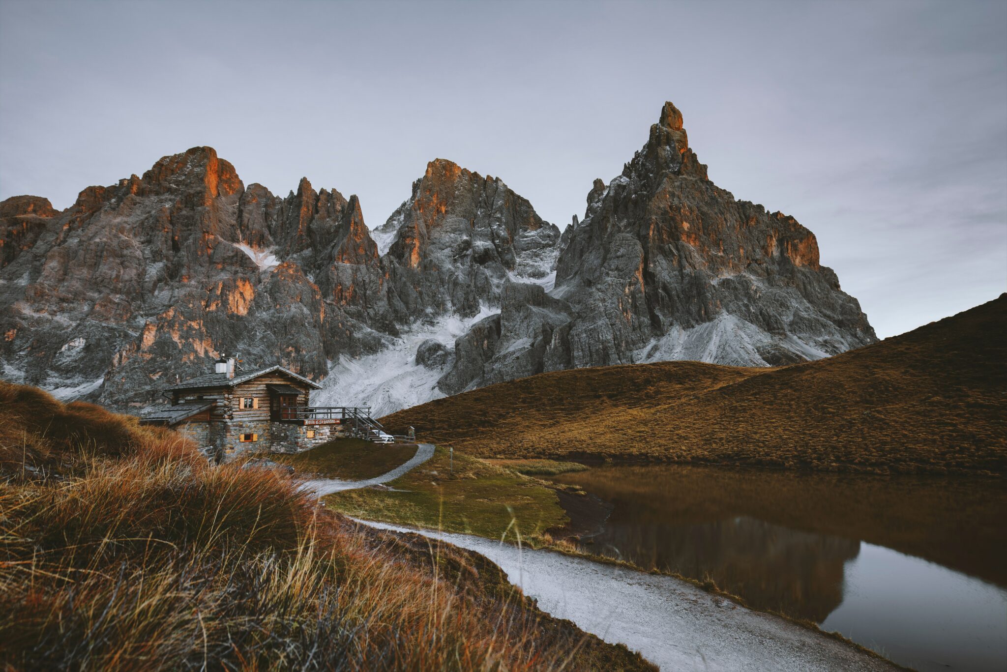 The image shows a mountain hut in the Dolomites, a beautiful range of mountains in northeastern Italy. The hut is situated in front of the impressive silhouette of Schlern Mountain in front of a clear sky. The evening sun reflects off the rocks.