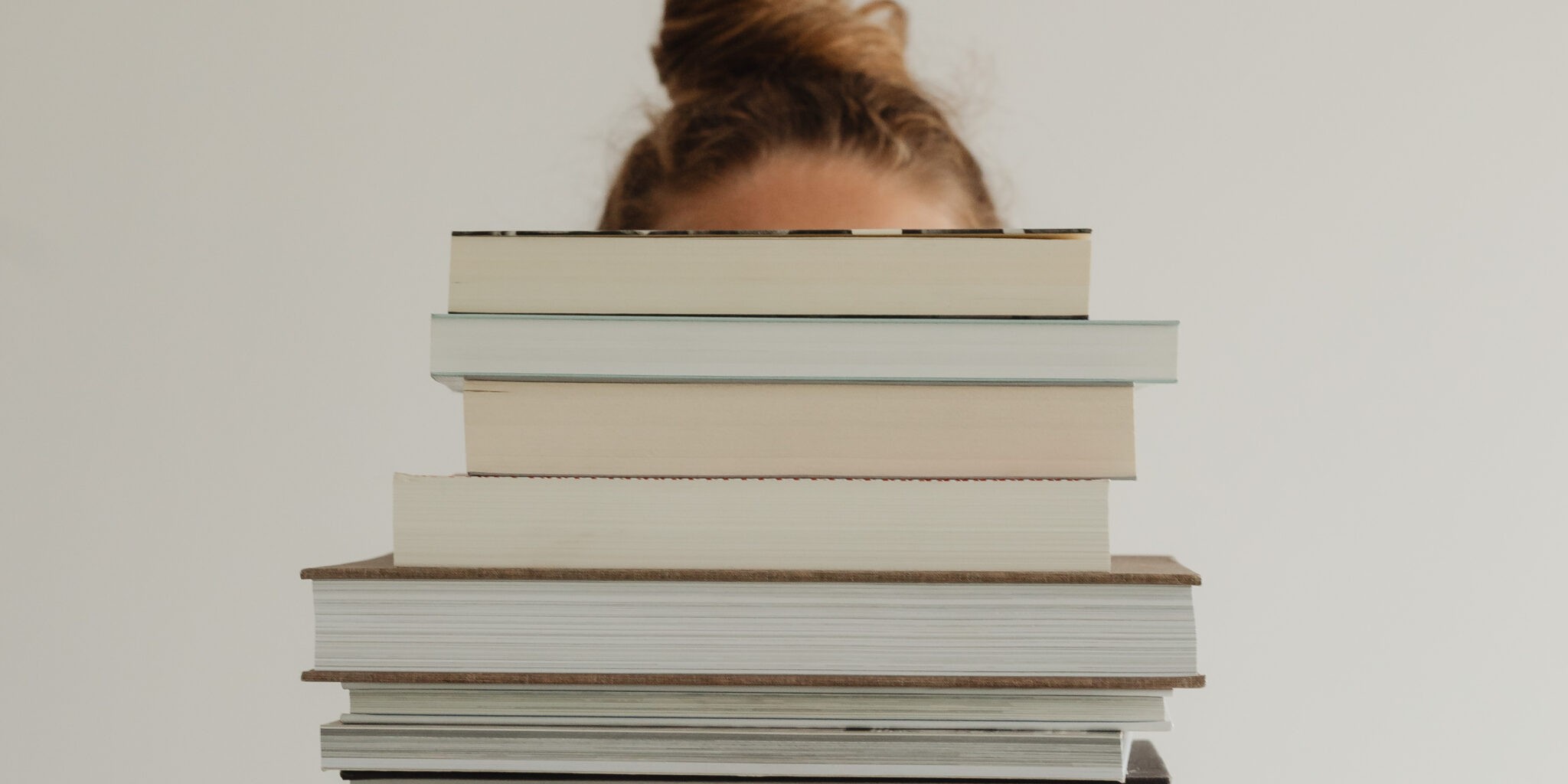 Image of a woman holding a pile of books covering her face. The picture is relevant to a blog post titled '4 Must-Reads That Go Beyond the Typical Branding Books'.