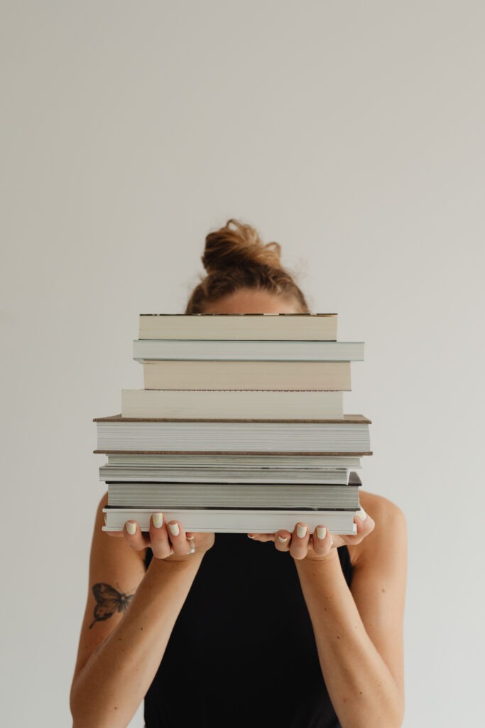 Image of a woman holding a pile of books covering her face. The picture is relevant to a blog post titled '4 Must-Reads That Go Beyond the Typical Branding Books'.