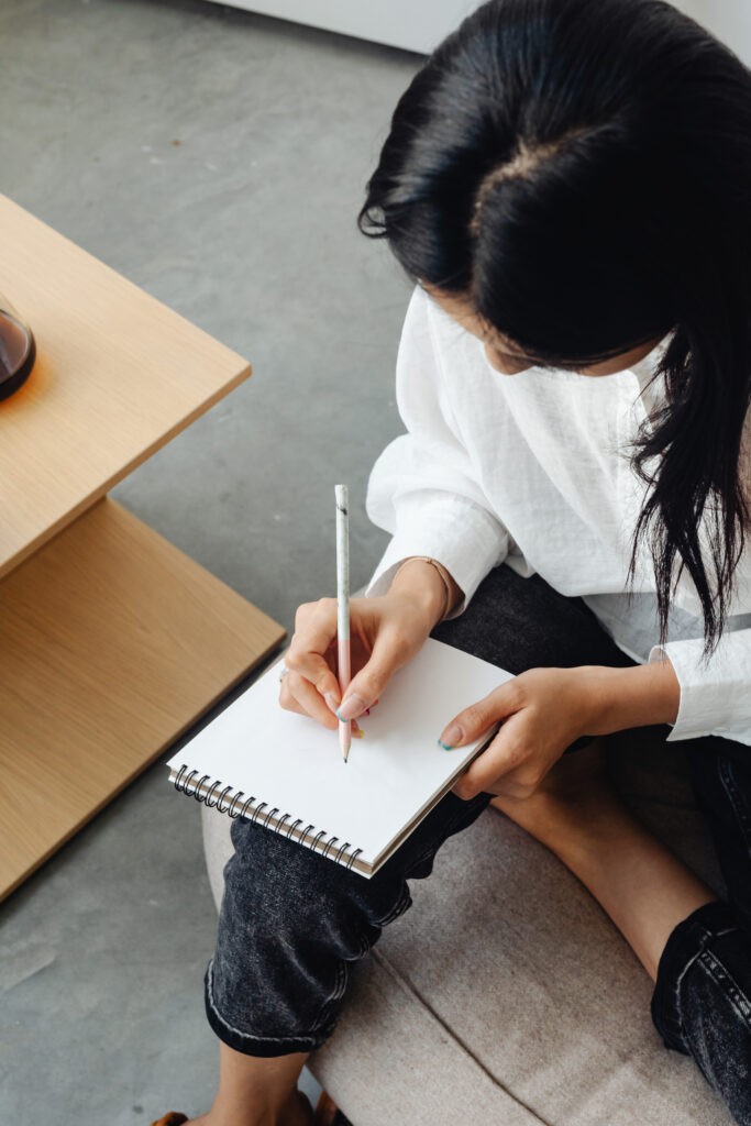 A woman sits with a pen in hand, poised above a blank notebook. The focus is on her hand and the notebook. The image is symbolic of the topic of brand voice, representing the blank canvas one faces when starting to develop their brand voice.