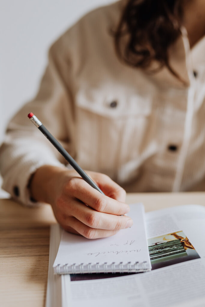 Das Bild einer Frauenhand, die einen Stift hält und schreibt, symbolisiert das Schreiben einer Tagline. Dieses Bild leitet den Blogbeitrag mit dem Titel "Was ist eine Tagline? Schritt-für-Schritt-Anleitung plus 50 Beispiele" ein.