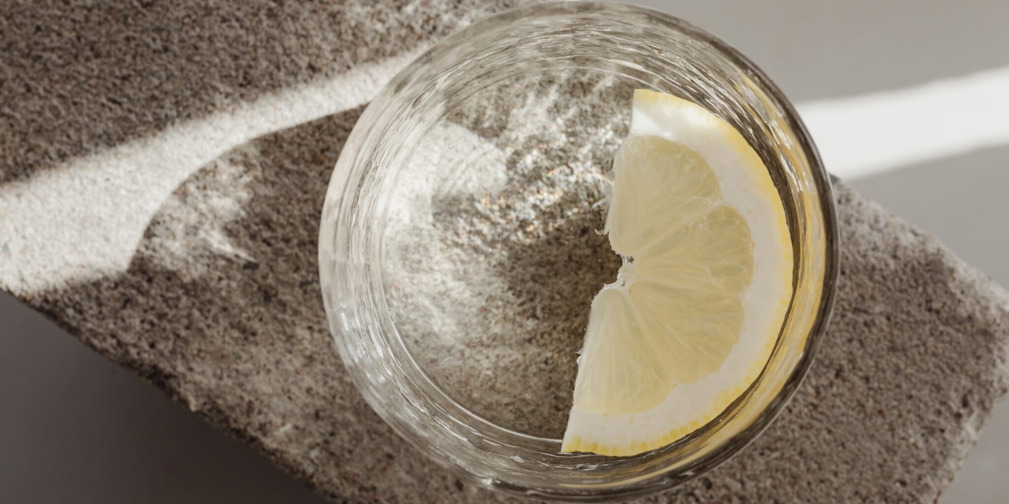 Top view of an unbranded glass of water with a lemon slice on a stone-textured surface against a light curtain background, conveying a brand-free, refreshing, and elegant vibe, as an intro to my article 'A deep dive into branding'