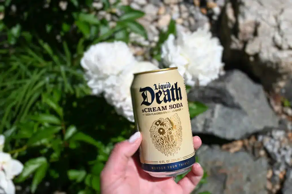 Eine Hand, die eine goldene Dose „Liquid Death Scream Soda“ vor grünem Laub und weißen Blüten hält, wobei die markante Heavy-Metal-Frakturschrift der Marke besonders hervorgehoben wird.