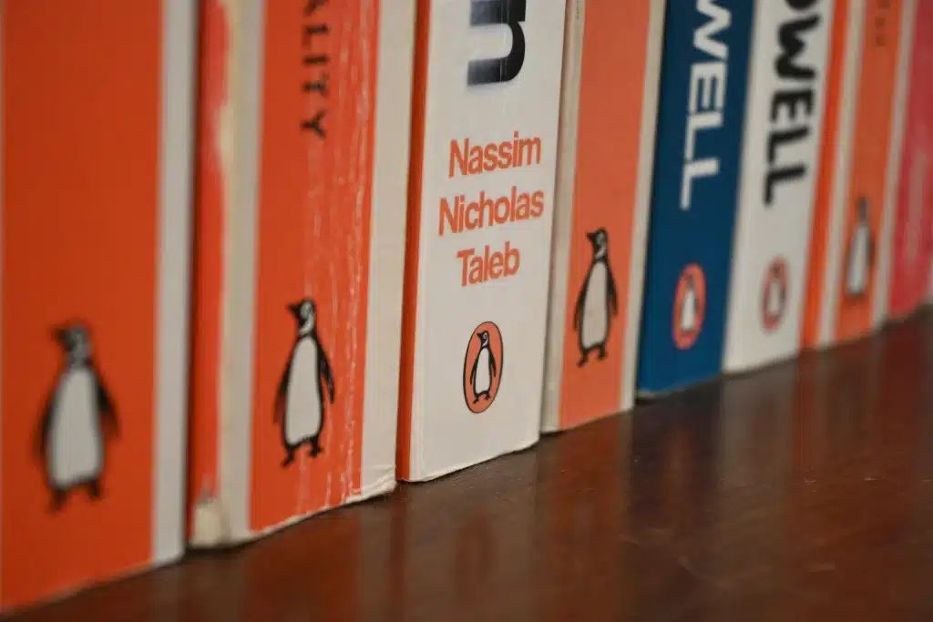 Eine Nahaufnahme mit geringer Schärfentiefe von Vintage-Penguin-Books-Buchrücken, aufgereiht auf einem dunklen Holzregal, die die ikonischen orange-weißen Farbbänder und das Pinguin-Bildmarkenlogo hervorhebt.