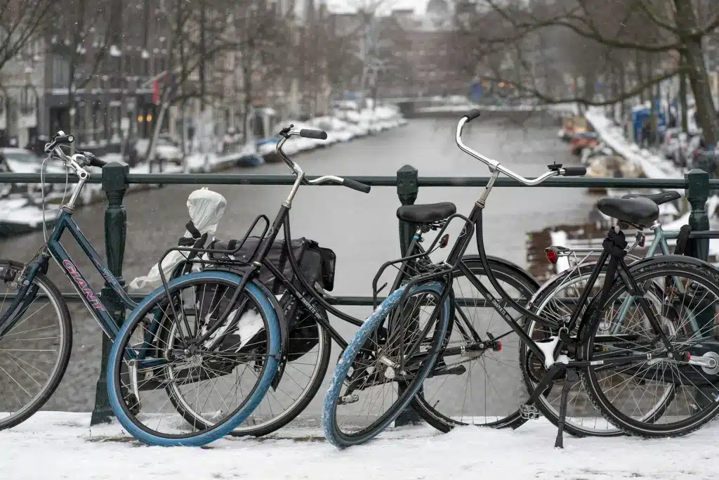Mehrere Citybikes an einem Metallgeländer neben einer Amsterdamer Gracht an einem verschneiten Tag, mit den unverwechselbaren leuchtend blauen Vorderreifen der Swapfiets-Leihräder.