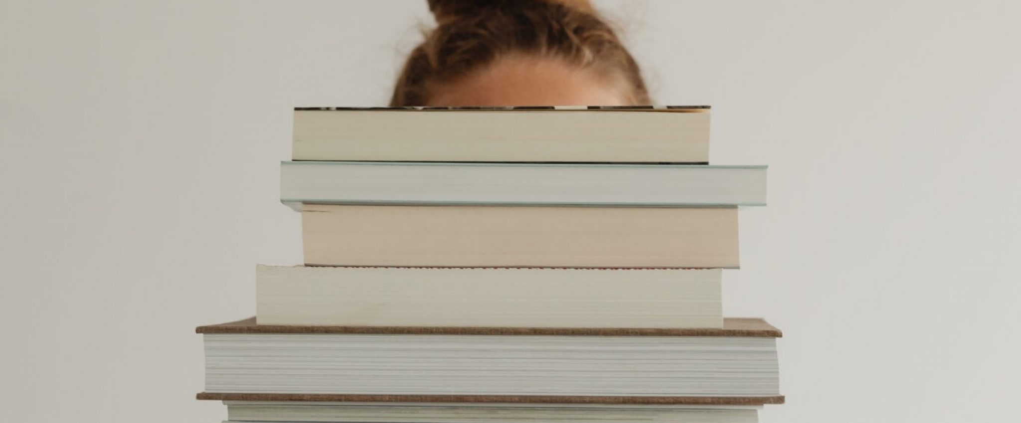 Image of a woman holding a pile of books covering her face. The picture is relevant to a blog post titled '4 Must-Reads That Go Beyond the Typical Branding Books'.