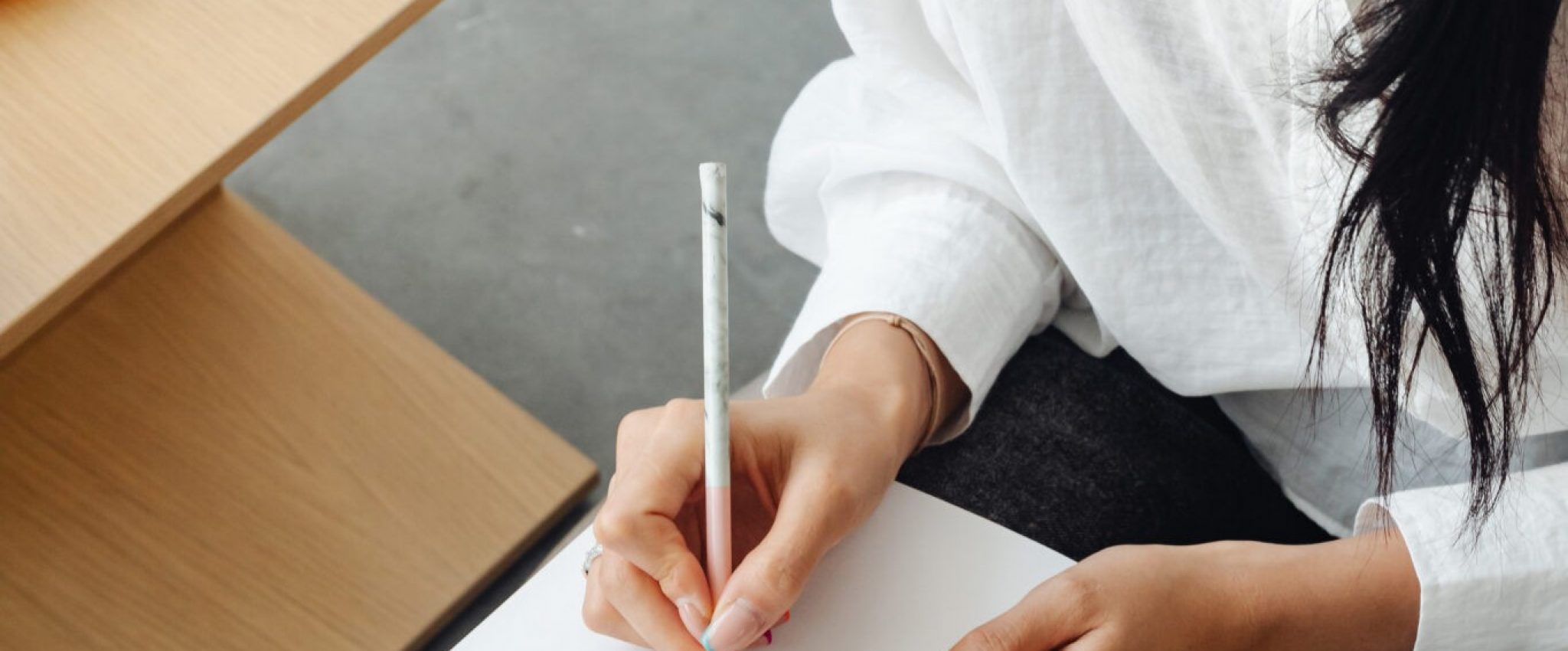 A woman sits with a pen in hand, poised above a blank notebook. The focus is on her hand and the notebook. The image is symbolic of the topic of brand voice, representing the blank canvas one faces when starting to develop their brand voice.